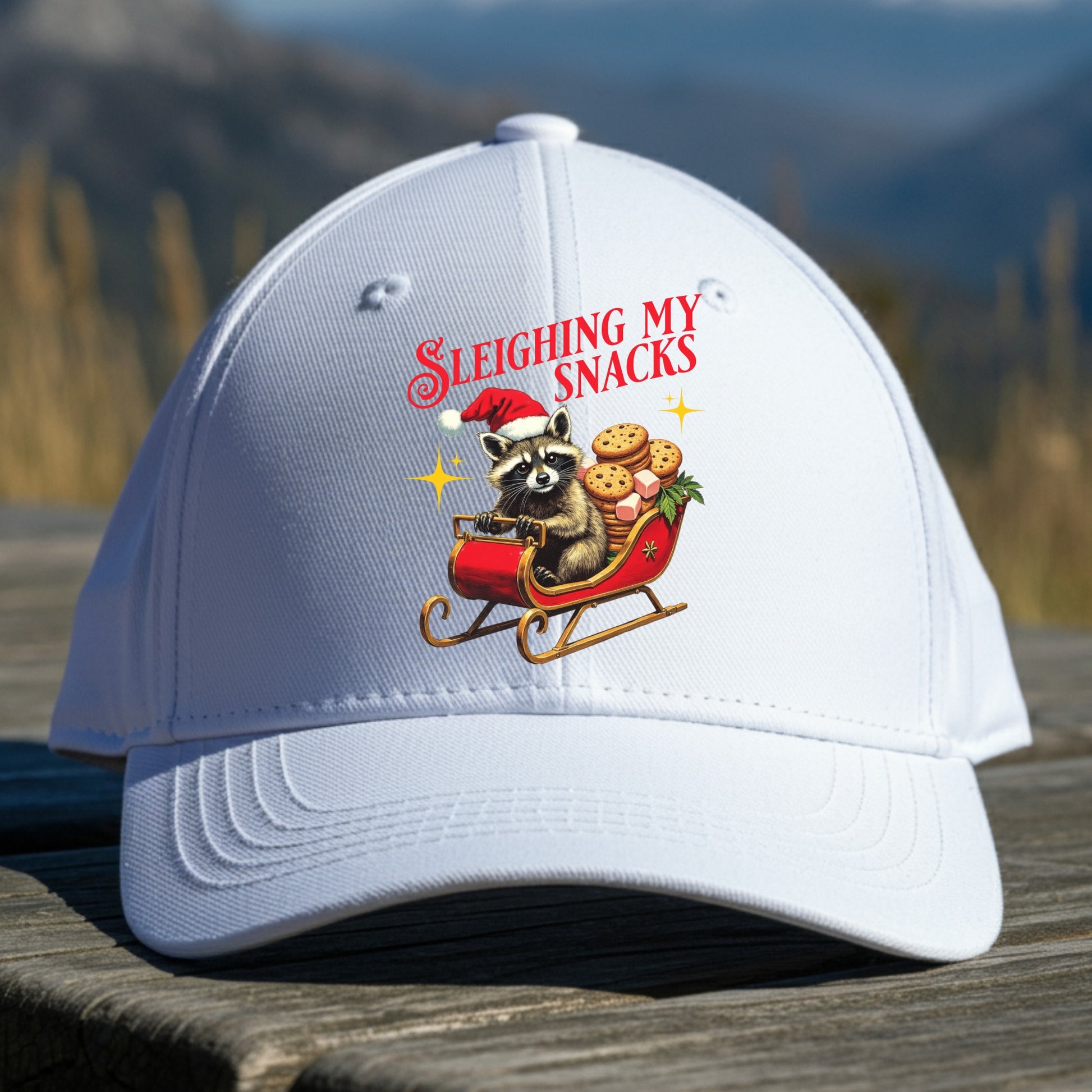 White cap with a festive design of a raccoon in a Santa hat with cookies, text 'Slething My Snacks', on a wooden surface with a mountain background.