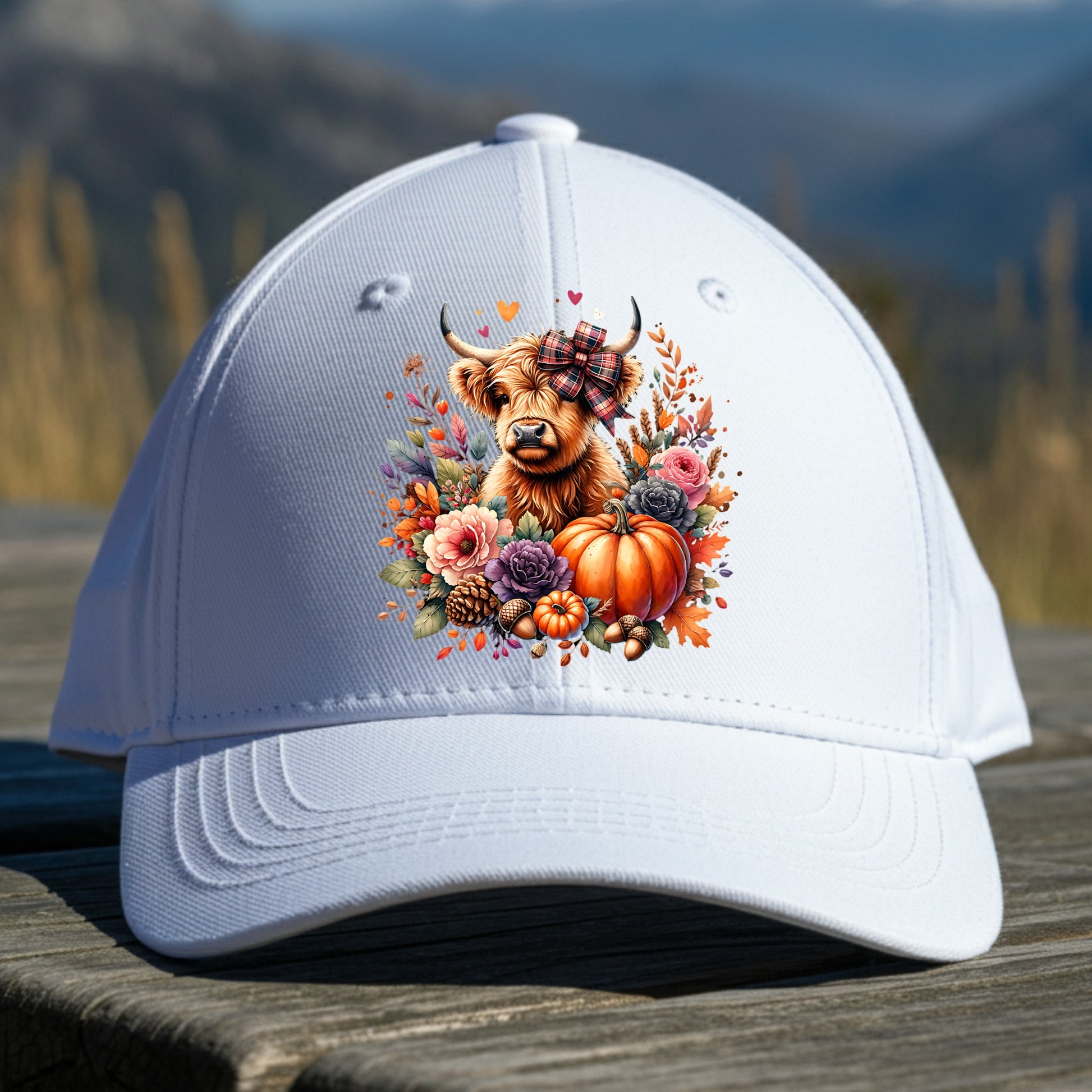 White cap with a decorative design of a Highland cow, flowers, and a pumpkin on a wooden surface with a mountain background.