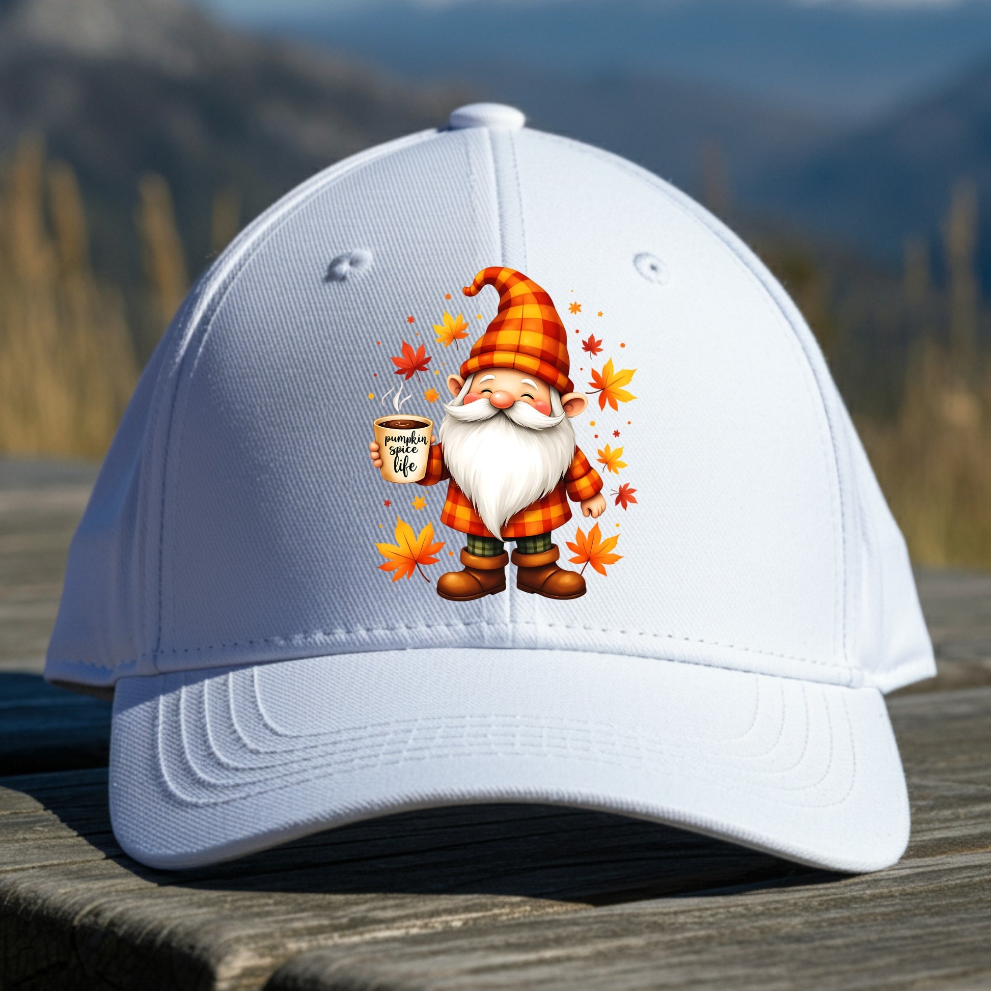 White cap with a cartoon gnome design featuring a mug and autumn leaves, placed on a wooden surface with a mountainous background.