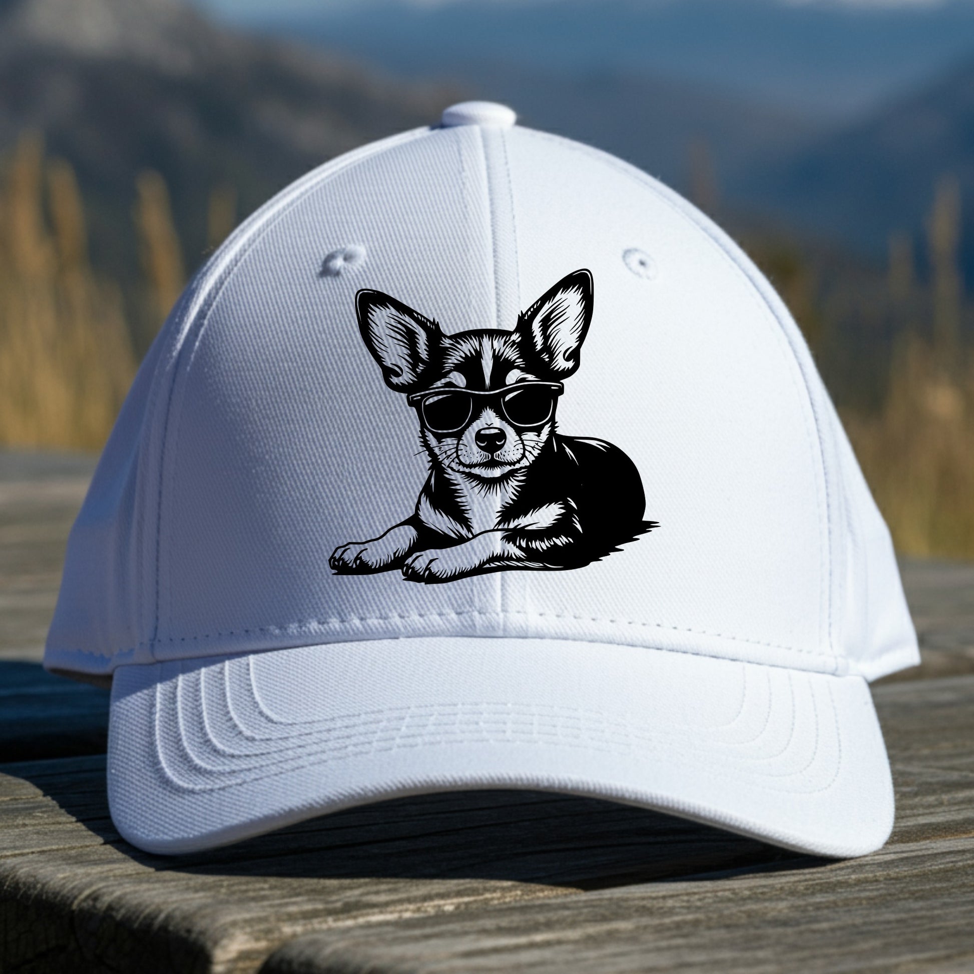 White cap with a chihuahua illustration wearing sunglasses on a wooden surface with a mountain background