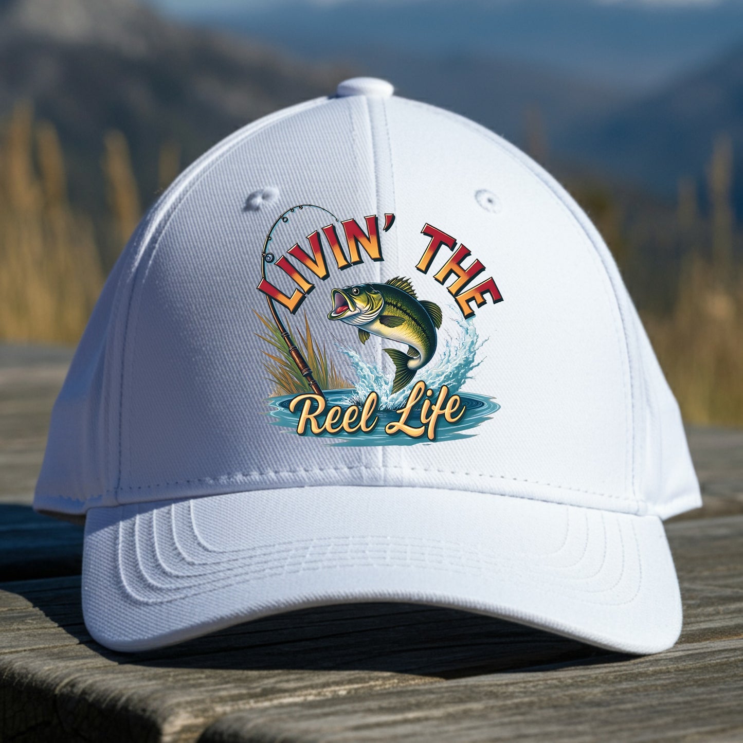 White cap with 'Livin' the Reel Life' fishing graphic on a wooden surface with a mountain background
