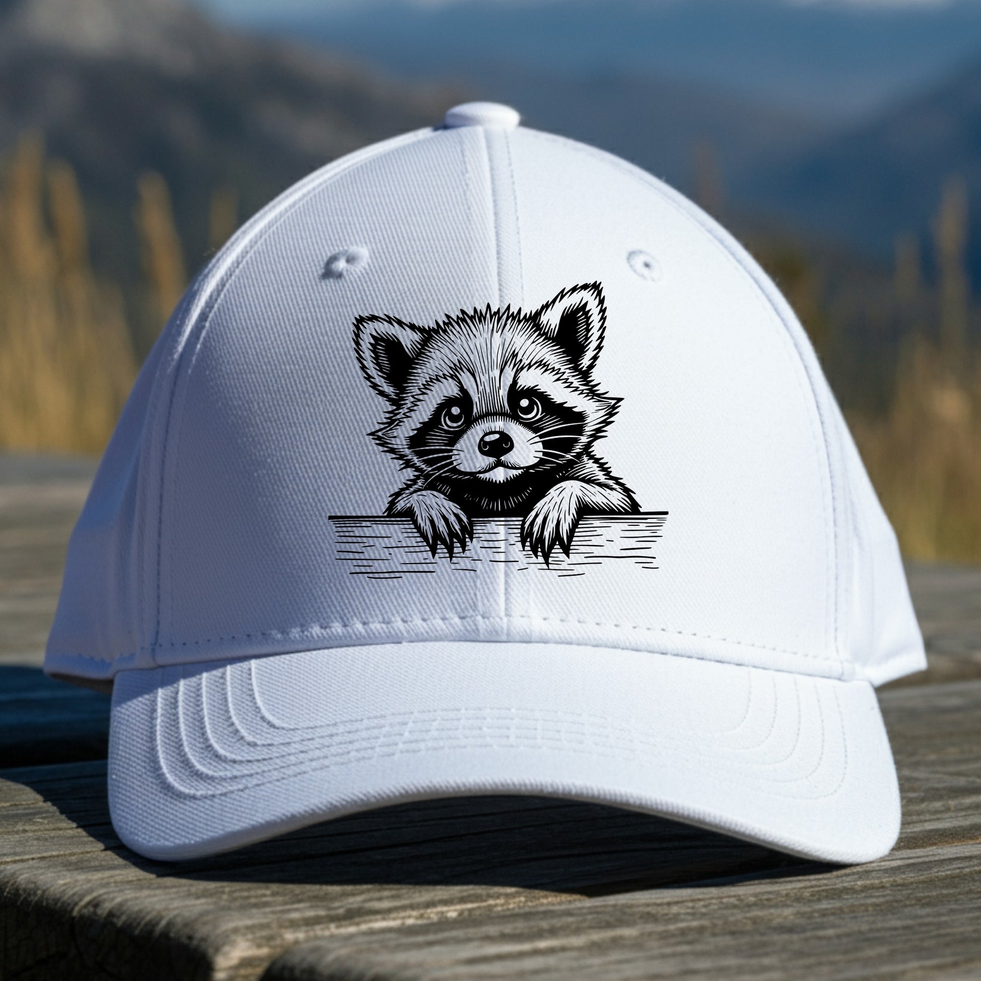 White cap with raccoon design on a sitting on a wooden surface with a mountain background
