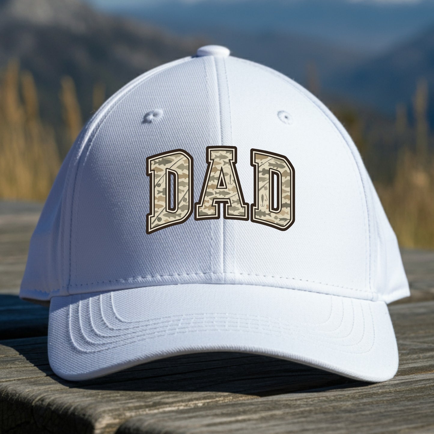White cap with 'DAD' embroidered on it, placed on a wooden surface with a mountainous background.