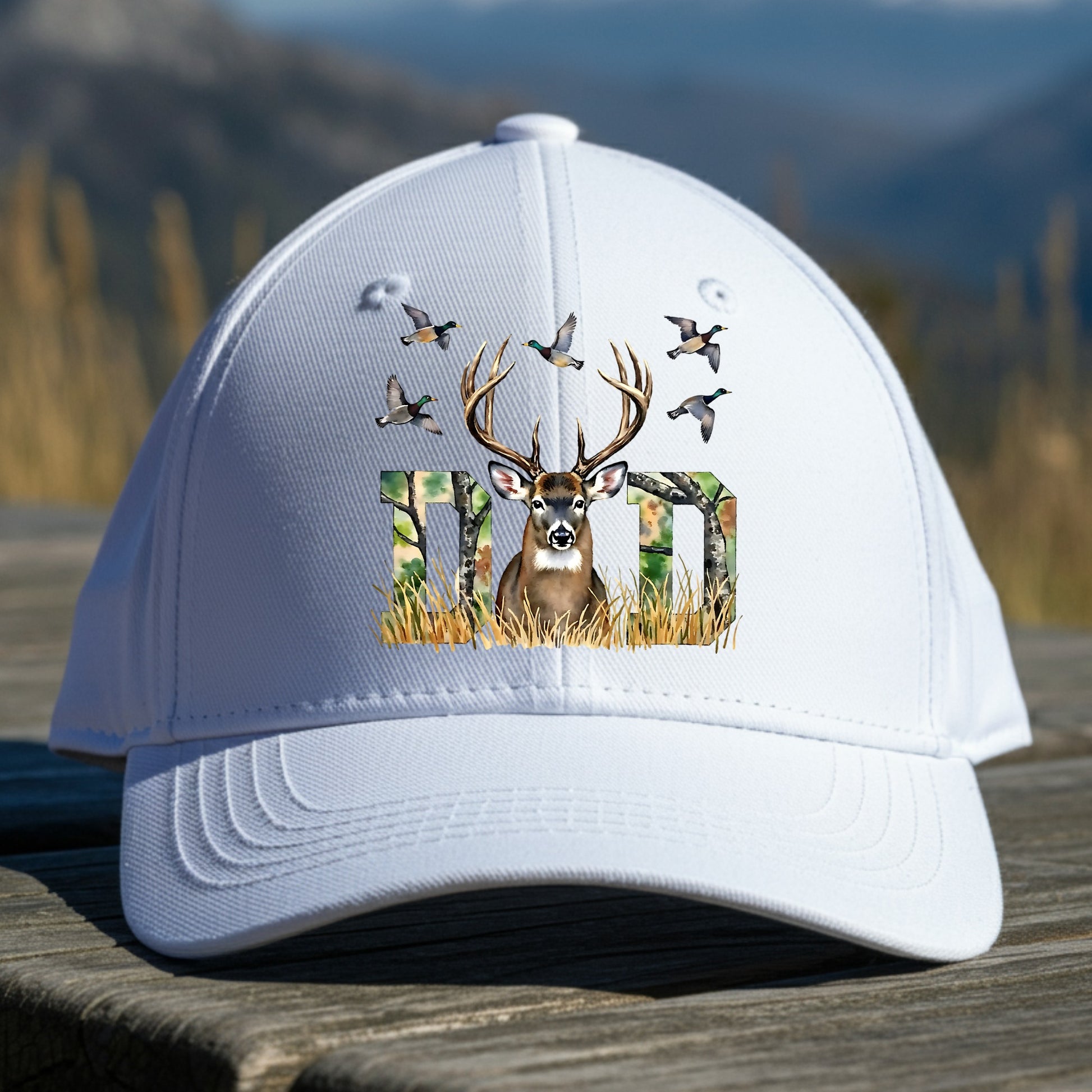 White cap with deer and hunting-themed design on a wooden surface with a mountain background