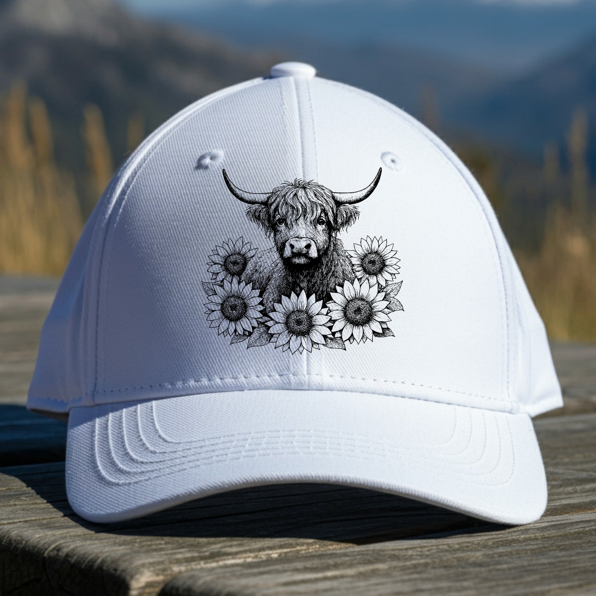 White cap with a black and white design of a cow and flowers on a wooden surface with a mountain background