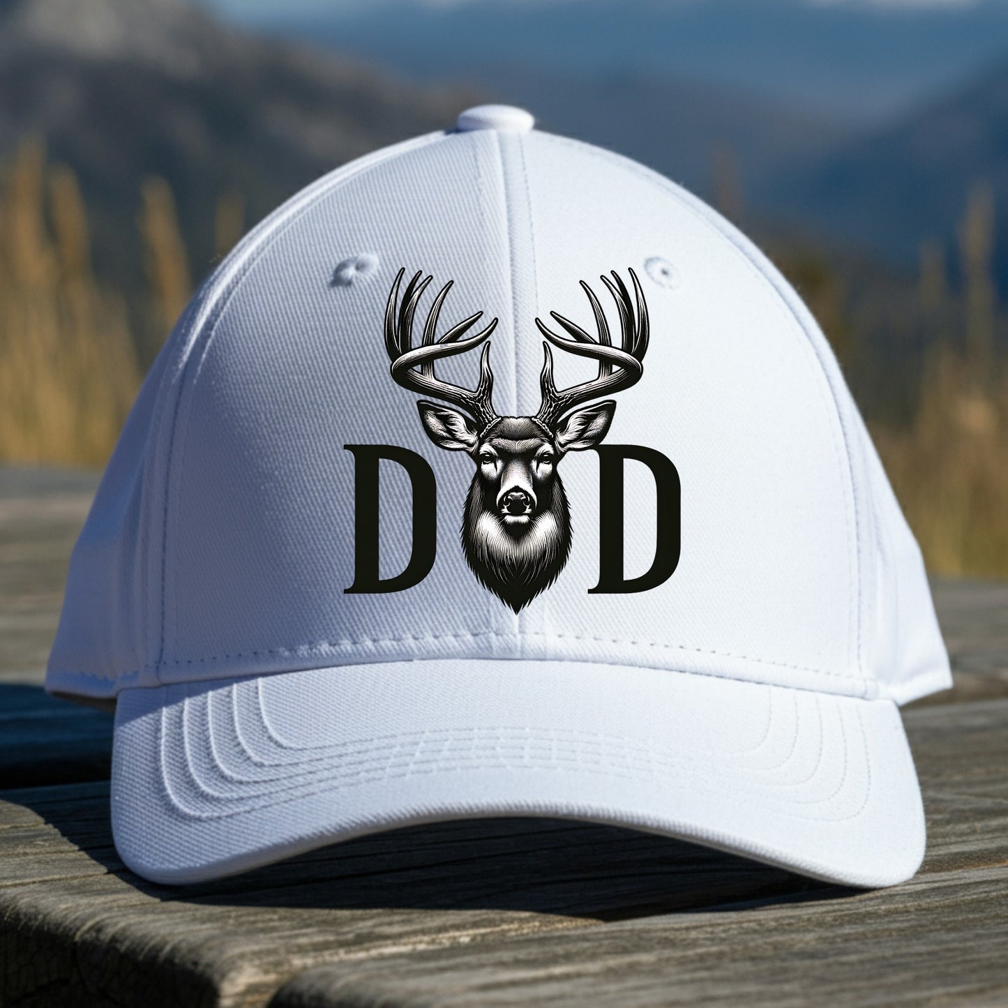 White cap with deer design and 'DAD' text on a wooden surface with a mountain background