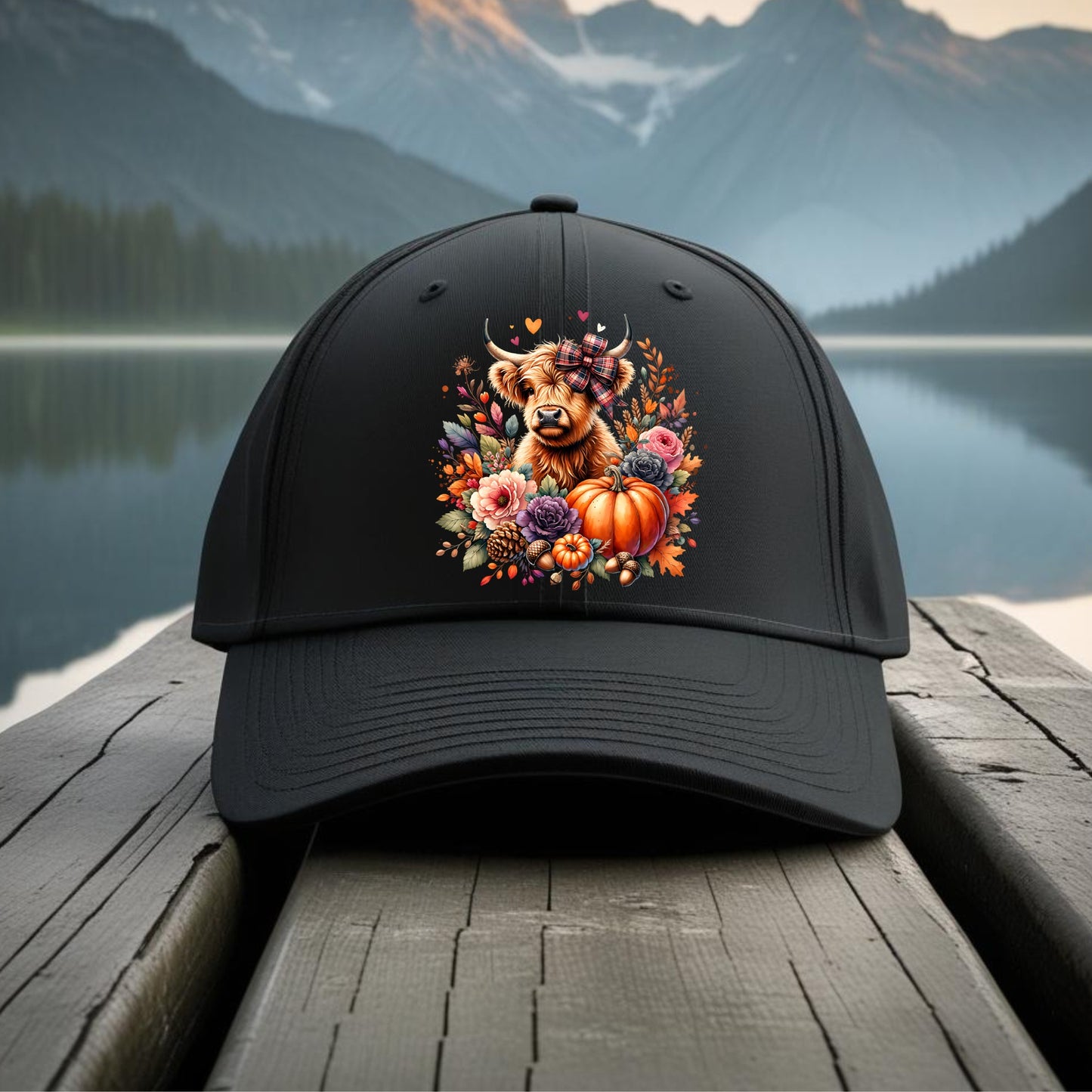 Black cap with a cow and pumpkin design on a wooden dock with a mountain and lake background