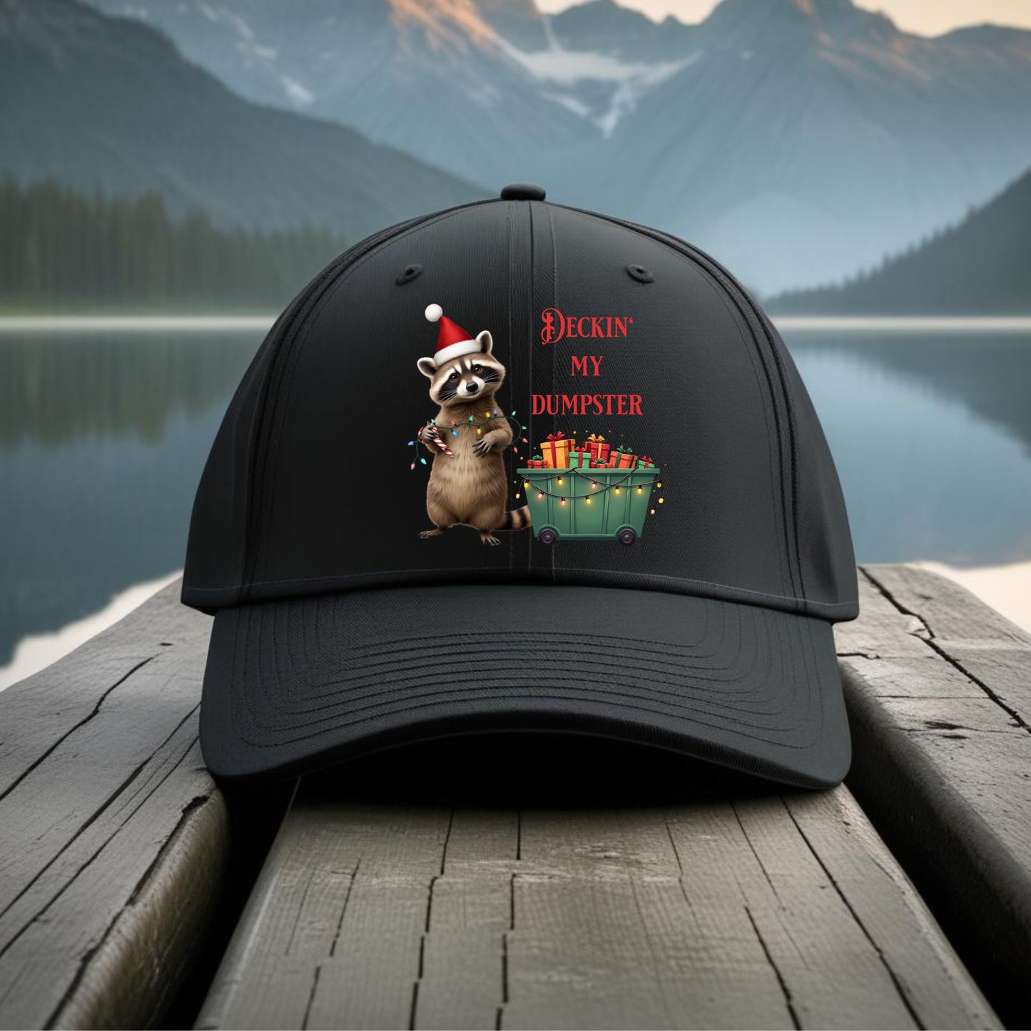 Black cap with a raccoon design and text 'Deckin' My Dumpster' on a wooden dock with a mountain and lake background.