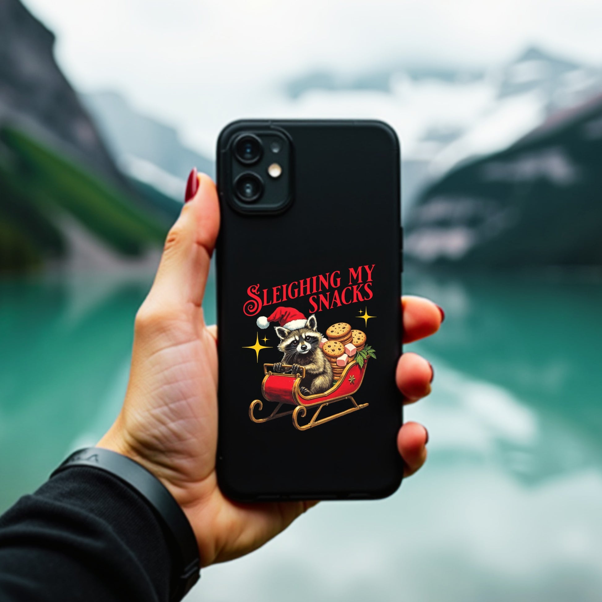 Hand holding a phone with a Christmas-themed case featuring a raccoon and text, against a blurred mountainous background.