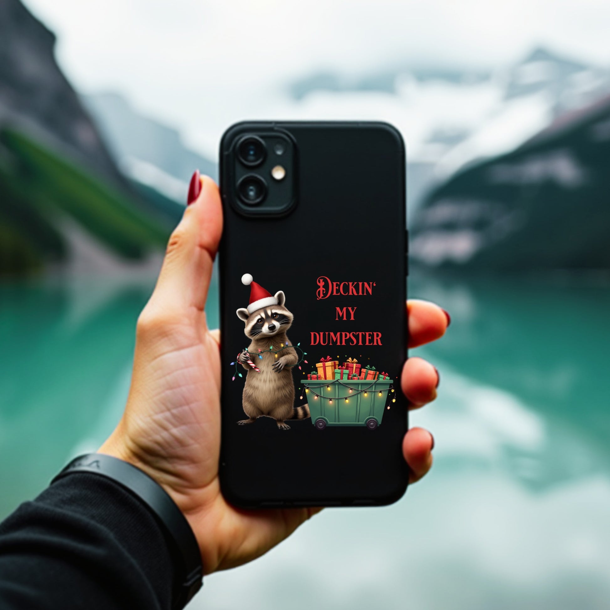 Hand holding a phone with a Christmas-themed case featuring a raccoon and text, against a blurred natural background.