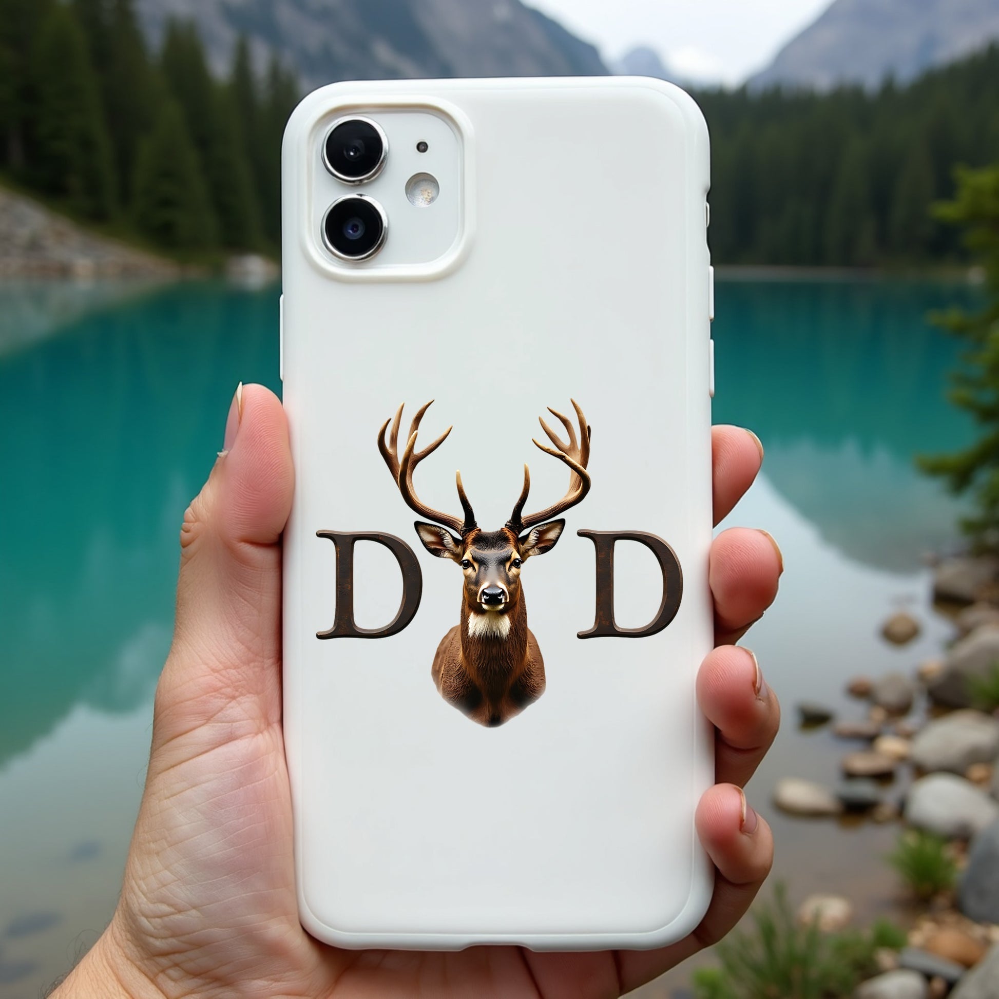 Phone case with deer design and 'DAD' text held in front of a scenic lake and mountains.