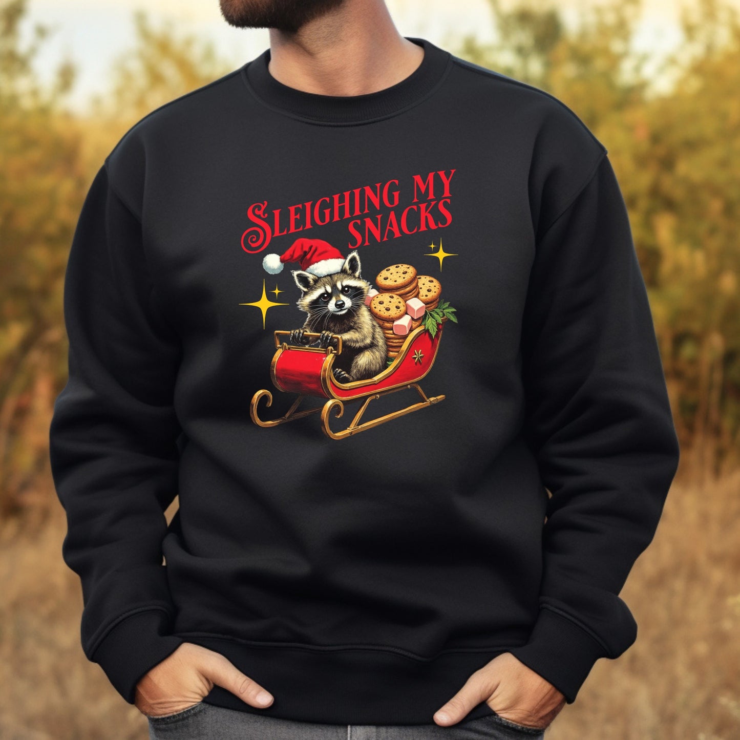 Black sweatshirt with a festive design of a raccoon in a sleigh with cookies, text 'Sleighbing My Snacks', against a blurred natural background.