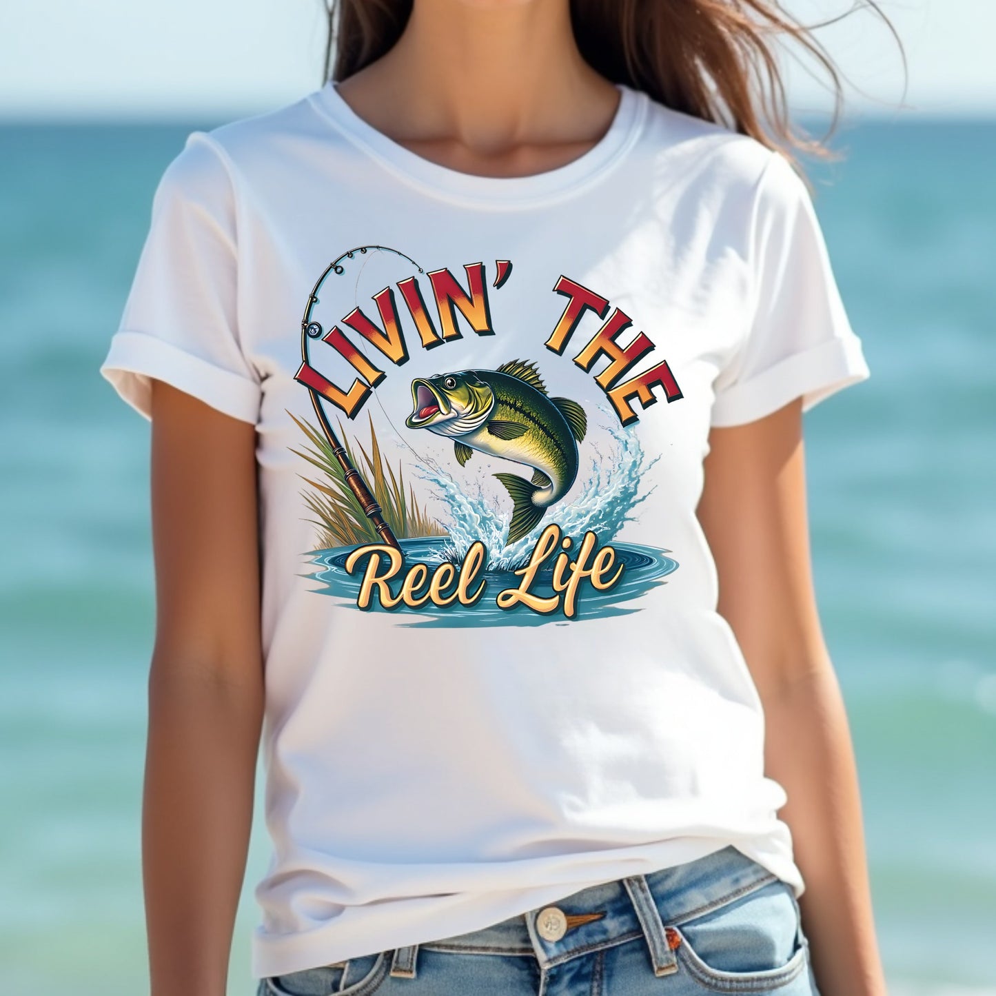 Woman wearing a white t-shirt with 'Livin' the Reel Life' graphic design by the ocean.