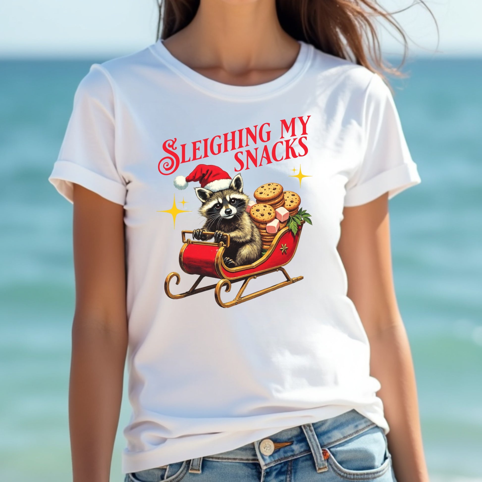 Woman wearing a white t-shirt with a Christmas-themed design of a raccoon sledding with cookies, against a blurred beach background.