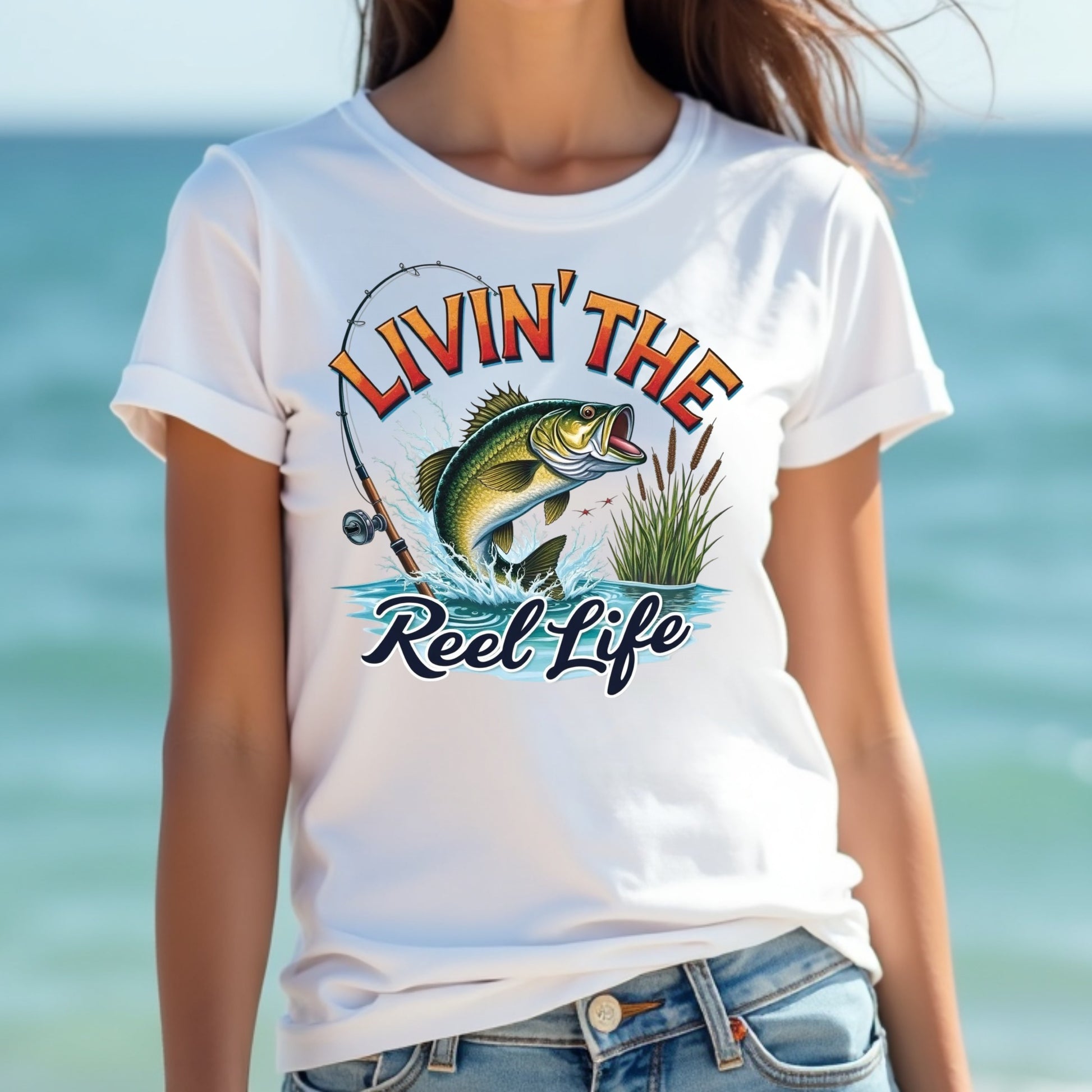 Woman wearing a white t-shirt with a fishing graphic and text 'Livin' The Reel Life' against a blurred beach background.