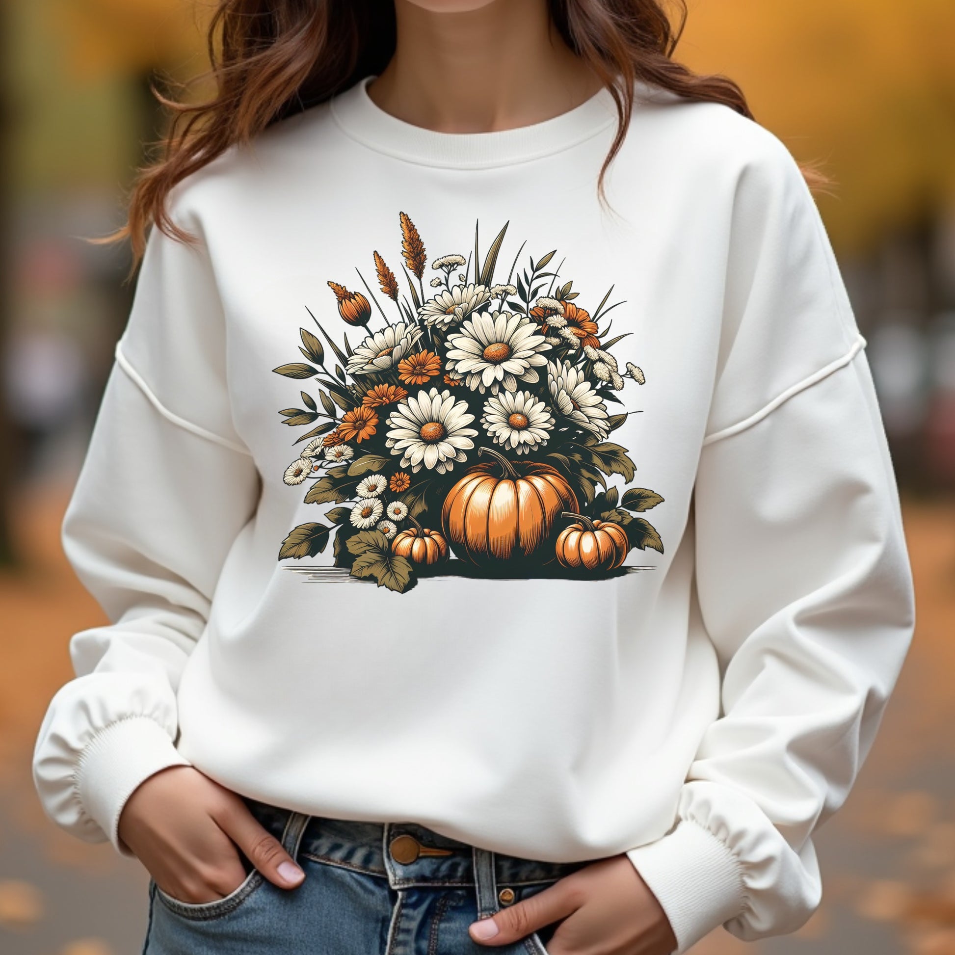 White sweatshirt with autumn-themed design featuring pumpkins and flowers, worn by a person outdoors.