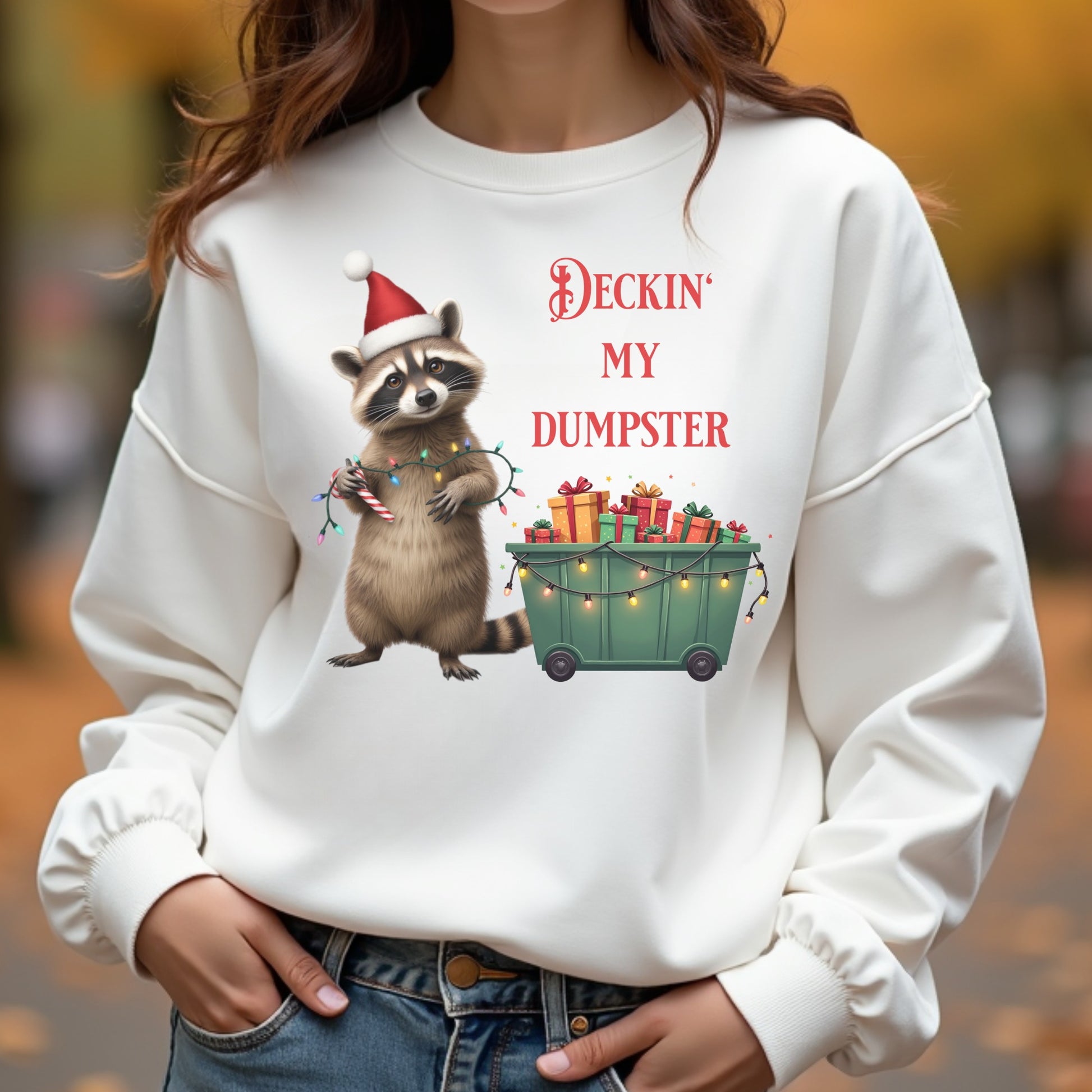 White sweatshirt with a raccoon wearing a Santa hat and Christmas lights design, text 'Deckin' My Dumpster', on a blurred autumn background.