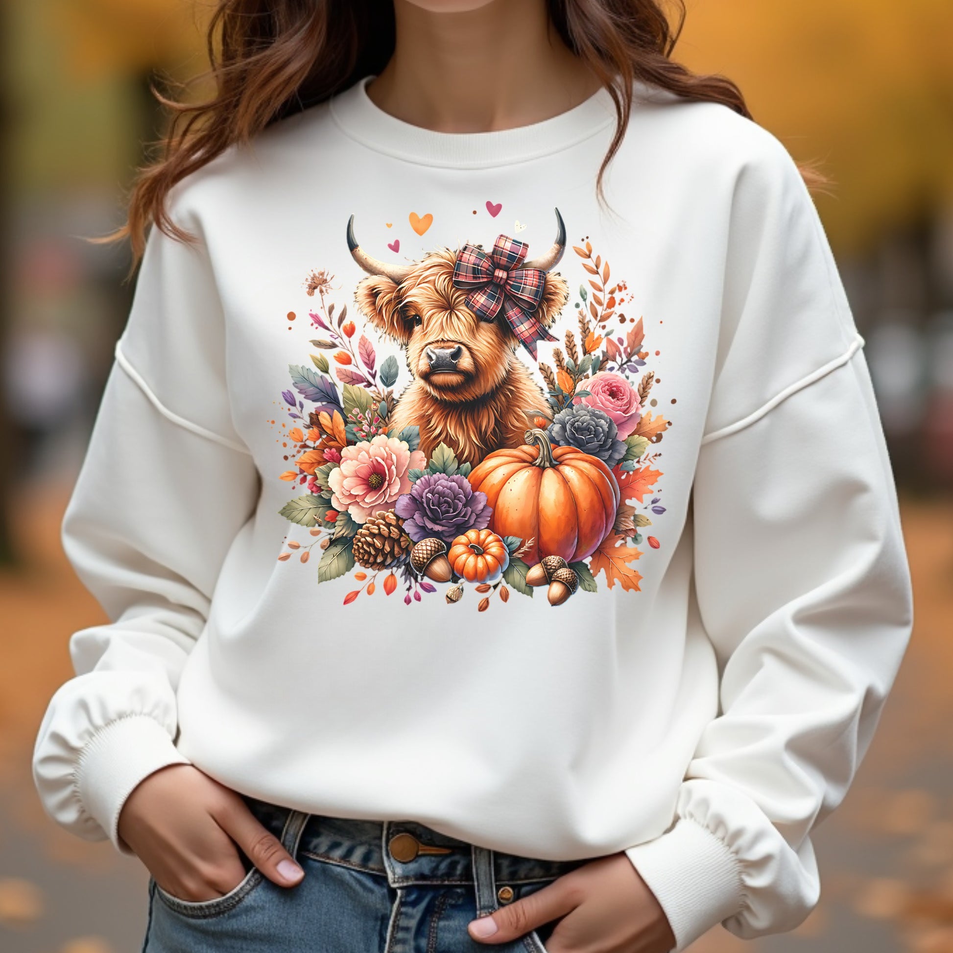 White sweatshirt with a highland cow and autumnal design worn by a woman outdoors.
