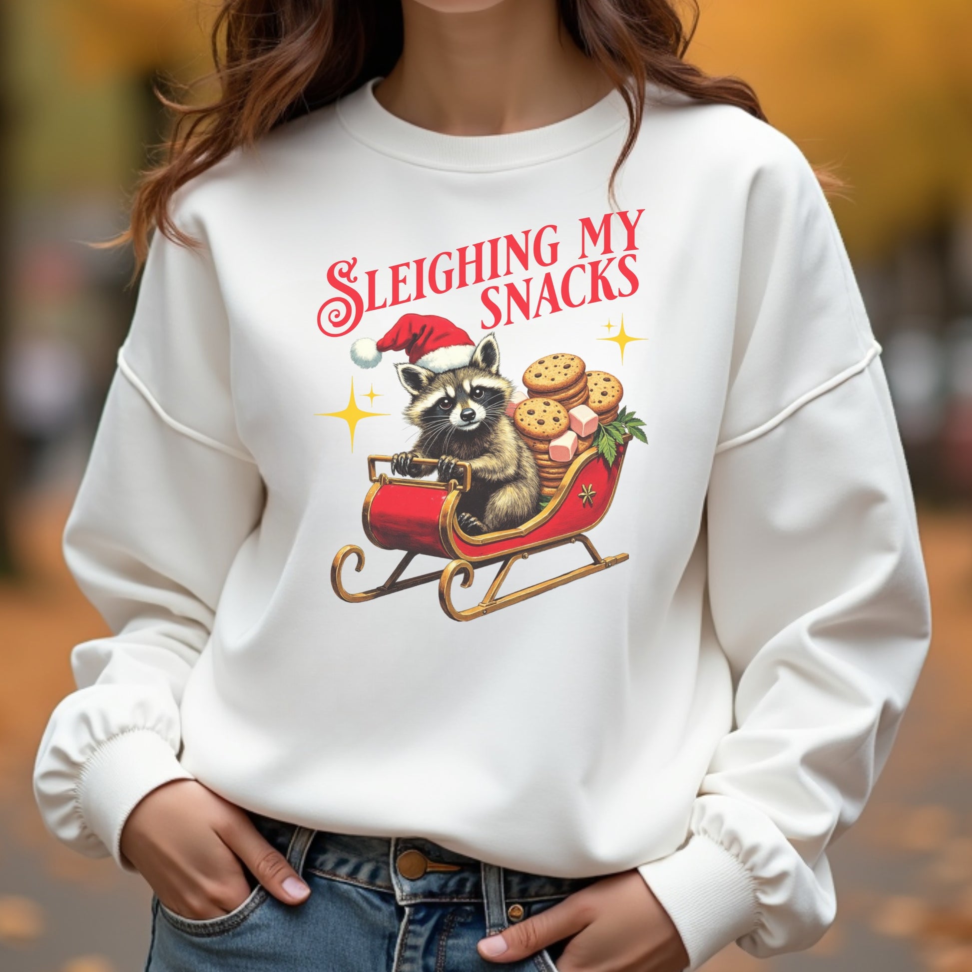 White sweatshirt with a festive design of a raccoon in a sleigh with cookies and text 'Sleighing My Snacks' worn by a woman outdoors.