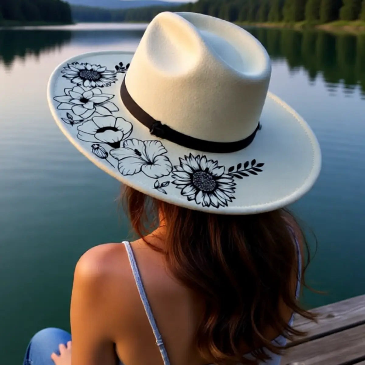 A woman sitting by a lake wearing a decorative sunflower design hat.