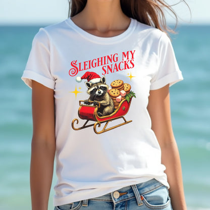 Woman wearing a white t-shirt with a Christmas-themed design of a raccoon sledding with cookies, against a blurred beach background.